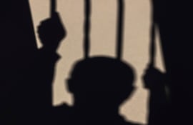 Silueta de un hombre con dos manos en barrotes, proyectando sombras sobre una superficie clara.