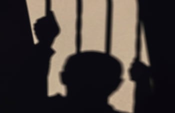 Silueta de un hombre con dos manos en barrotes, proyectando sombras sobre una superficie clara.