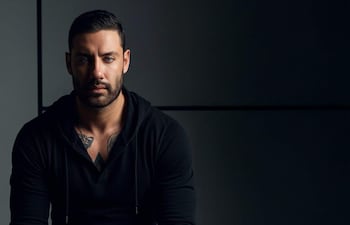 Daniel Dueñas sentado en un banquito con sudadera negra y pantalones oscuros, mostrando tatuajes en los brazos y expresión tranquila.