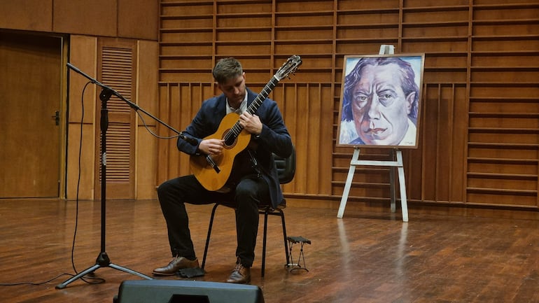 Músico en traje oscuro tocando guitarra en un auditorio con retrato de Mangoré al fondo.