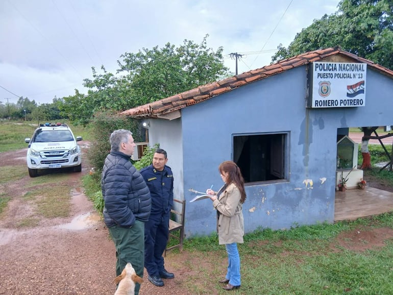 Hombre de chaqueta negra y pantalones oscuros conversan con mujer que toma notas, frente a la estación policial con cielo nublado.