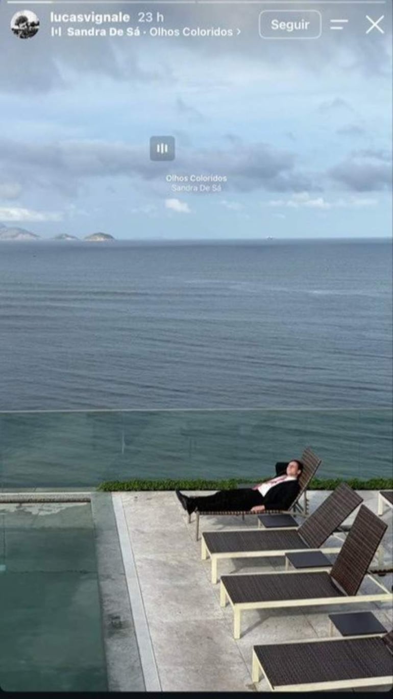Gaspi en traje oscuro y camisa clara reclinado en una silla, frente a un paisaje de mar y horizonte lejano.