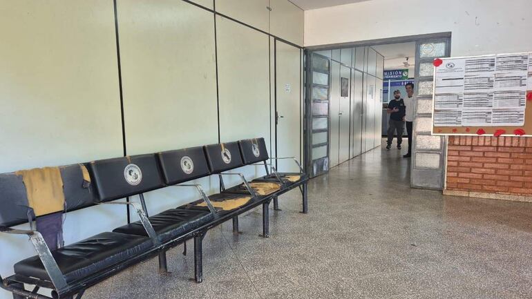 Dos hombres conversan; uno lleva camisa blanca y otro camiseta negra. Banco de espera desgastado en un ambiente austero.