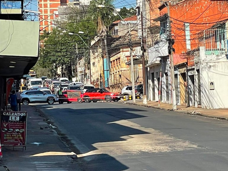 Escena urbana sin personas, con varios autos en movimiento y estacionados en una calle comercial cercana a tiendas de calzado.
