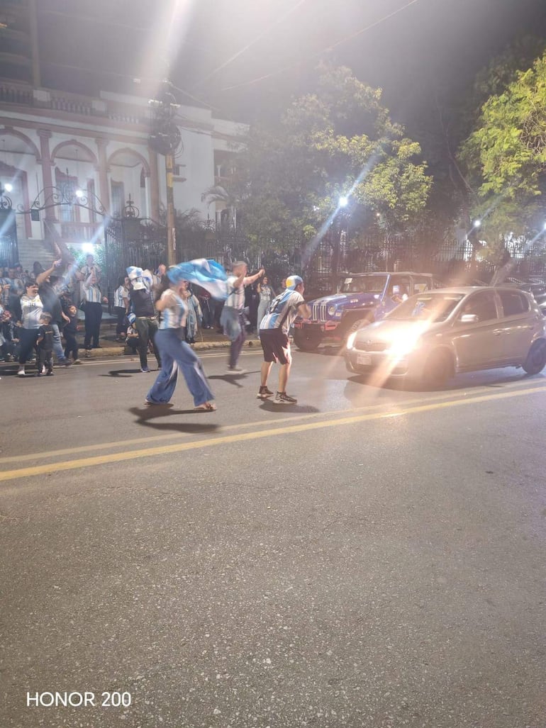 Personas celebrando en la calle con camisetas celeste y blancas, una persona levanta una bandera argentina en ambiente festivo nocturno.