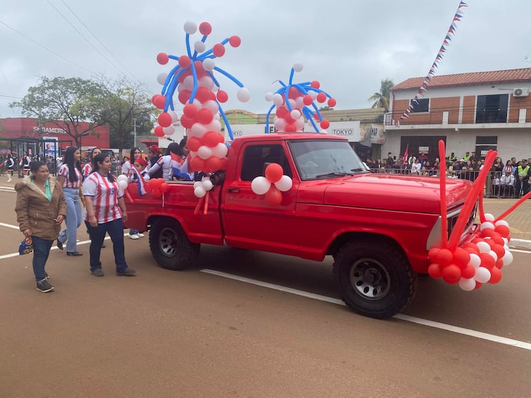 Cinco personas en camisetas rojas y blancas caminan junto a una camioneta decorada con globos en un festivo ambiente de desfile.