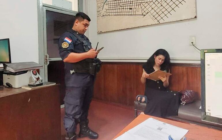 Mujer con vestido negro sentada en sofá, lectura de hoja, mientras oficial de policía supervisa con teléfono móvil en mano.