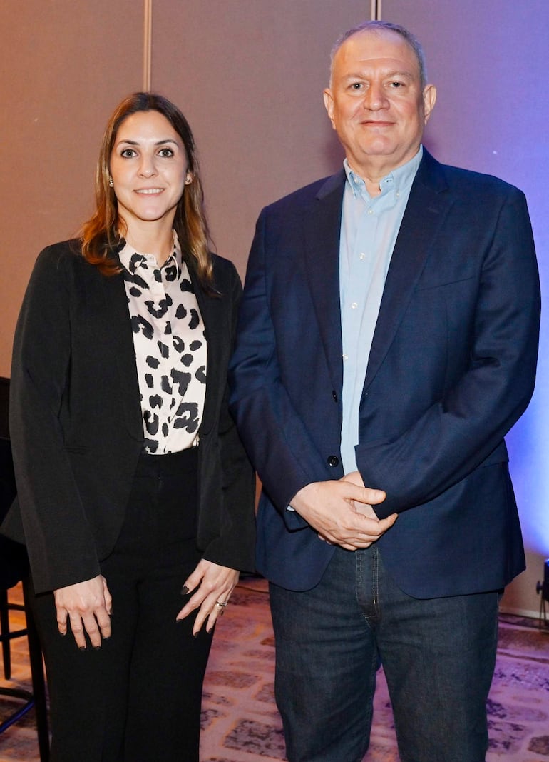 Mujer con blazer negro y blusa de manchas sonríe junto a hombre en camisa clara y saco oscuro en interior iluminado.