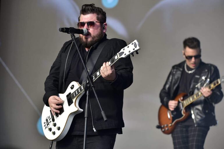 Músicos de Antenna en el escenario, uno canta con micrófono y guitarra blanca, otro toca guitarra marrón, ambos con gafas oscuras y chaquetas de cuero.