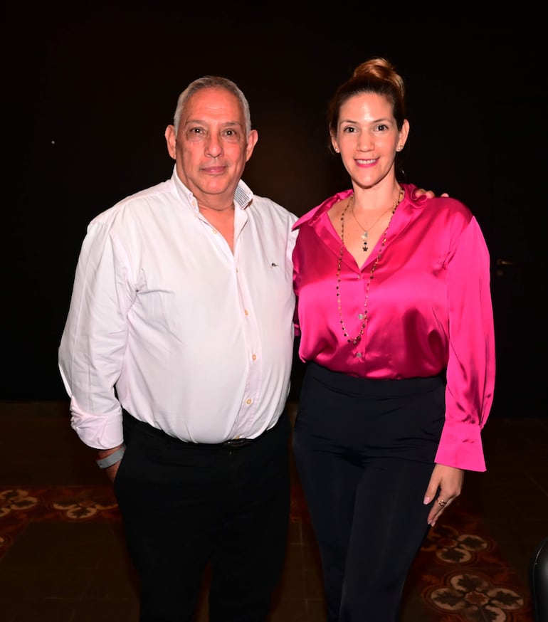 Un hombre canoso en camisa blanca y una mujer con blusa rosa conversan en un ambiente oscuro, ambos sonrientes.