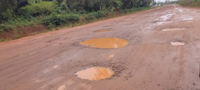 Carretera de tierra en mal estado con charcos de agua, rodeada de vegetación densa, sin personas visibles.