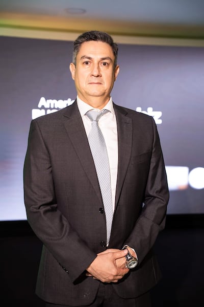 Carson Villalba en traje oscuro y corbata, de pie con expresión seria en un ambiente iluminado con fondo elegante.