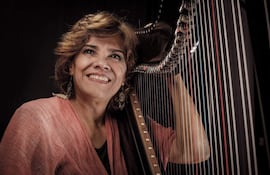Norma Ortega sonríe con cabello rizado y vestido rosa claro, sosteniendo un arpa en un fondo oscuro.