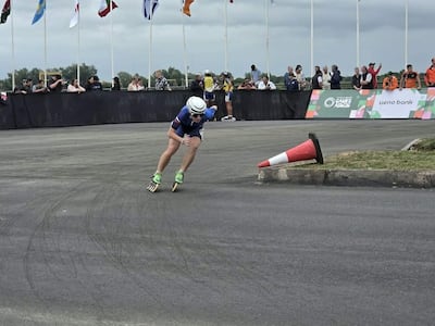 Patinador con traje azul y verde y casco blanco se desliza en carrera, mientras banderas ondean y espectadores observan atentos.