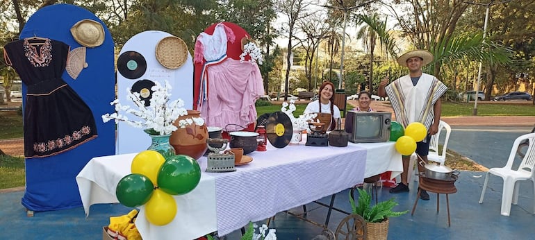 Hombre con poncho y sombrero sonríe a la derecha, mujer sentada en el centro y otra de pie a la izquierda, todos felices en un parque.