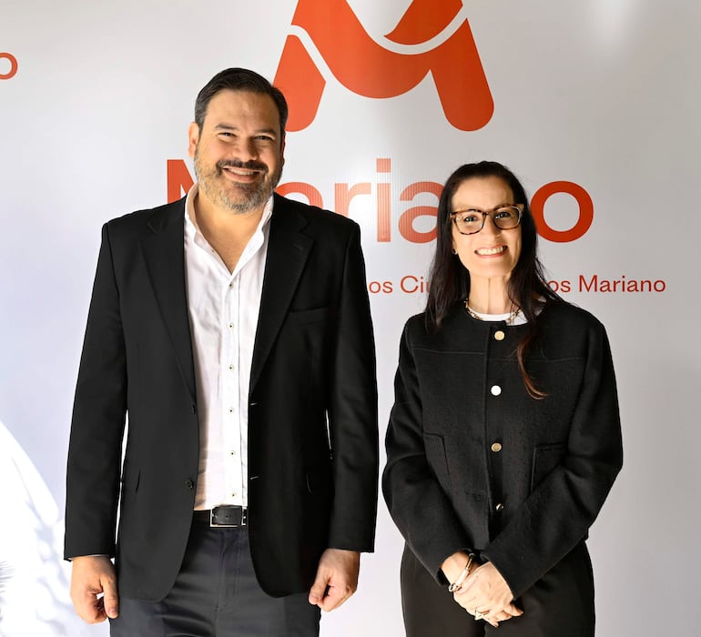 HOMBREsonríe con saco negro y camisa blanca junto a una mujer con chaqueta negra y gafas, frente al logo de Mariano.