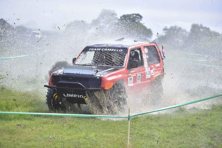 Vehículo off-road rojo del equipo TEAM CAMELLO en acción, levantando barro en una competencia de rally. Ambiente de campo difuso.