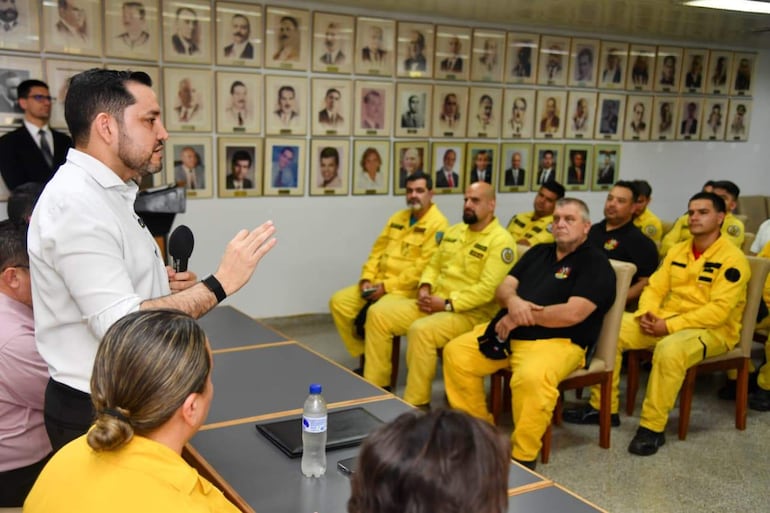 Hombre con camisa blanca habla sosteniendo un micrófono, rodeado de personas en uniformes amarillos, en un ambiente formal.