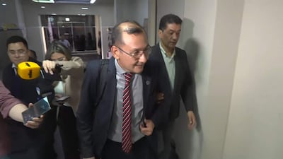 Miguel Mora, con corbata roja y camisa clara, se retira mientras una mujer lo entrevista, con un fondo de asistentes.