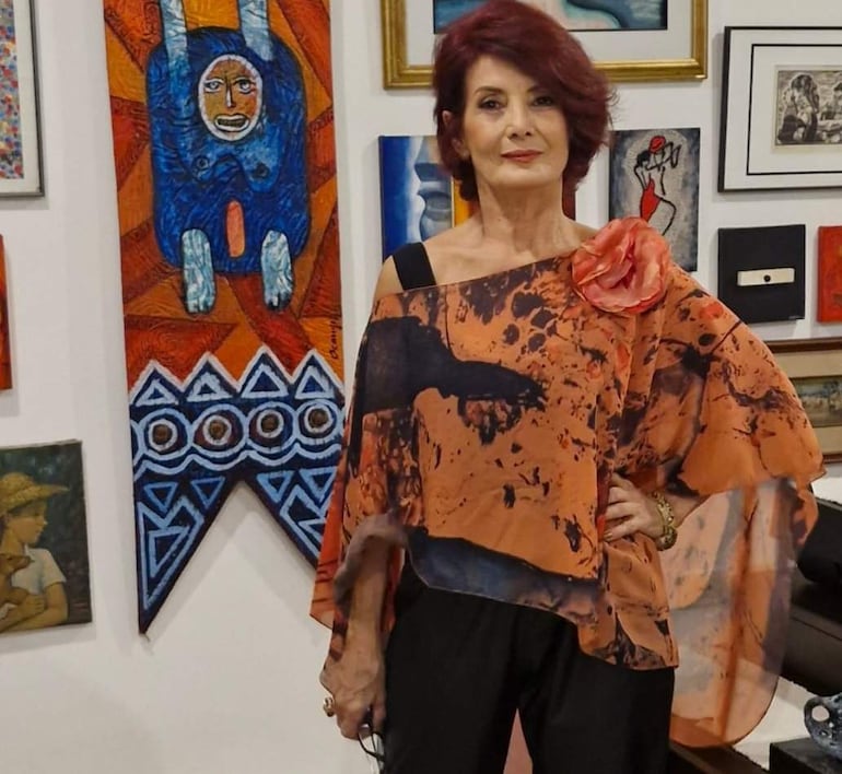 Mujer de cabello rojo con blusa artística en tonos naranjas y negros, posando con gafas en galería de arte decorada.