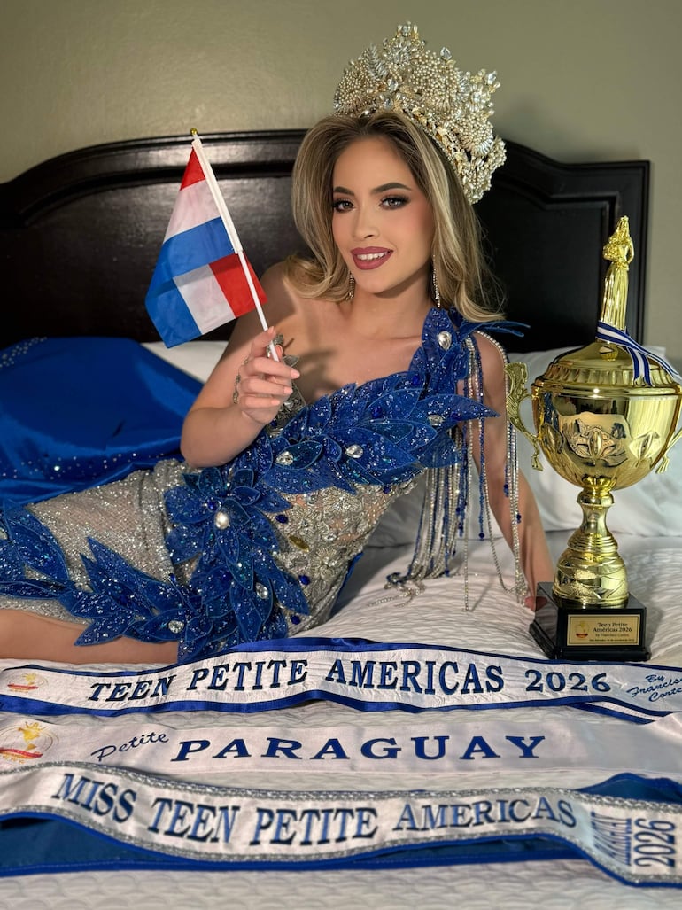 Joven mujer en traje de gala azul y plateado sonríe con trofeo dorado y bandera de Paraguay en un ambiente festivo.
