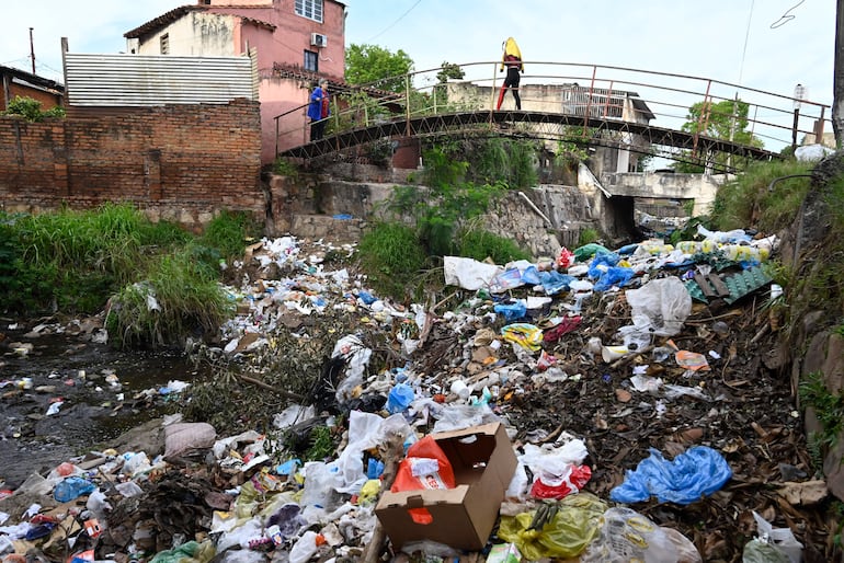 Una persona de pie en un puente observa un arroyo contaminado, mientras otro cruce con un atuendo oscuro y objeto amarillo en la cabeza.