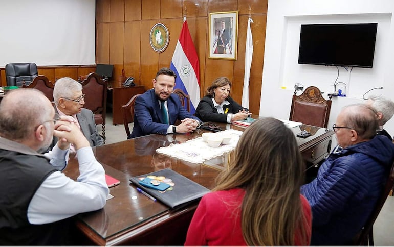 Derlis León, en camisa blanca y blazer oscuro, habla ante el Consejo del IPS en sala iluminada con bandera paraguaya de fondo.