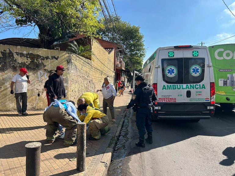 Personas con indumentaria de emergencia atienden a una estudiante en el suelo, cerca de una ambulancia en una calle urbana.