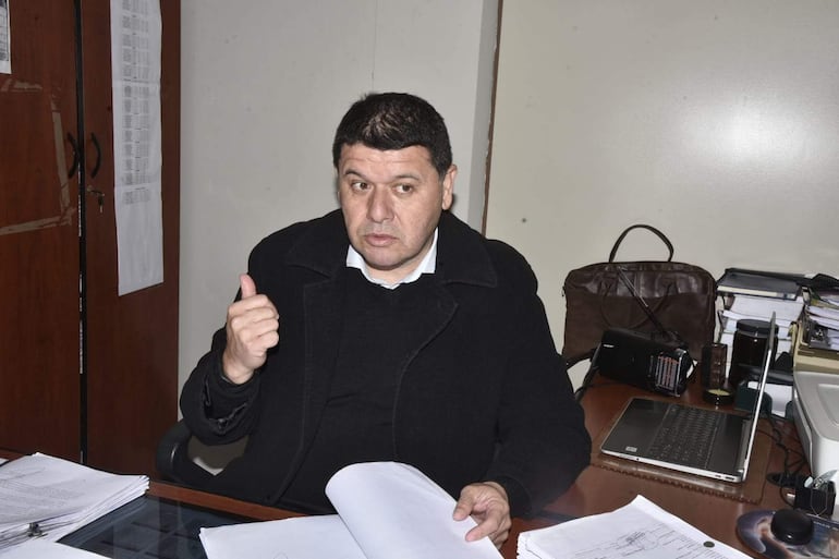 Eugenio Ocampos, vestido con abrigo negro y camisa blanca, gesticula en su oficina rodeado de documentos y una computadora portátil.