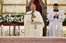 Clérigo de avanzada edad en vestimenta litúrgica blanca habla frente a un micrófono, con joven vestido de túnica blanca a su lado.