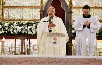 Clérigo de avanzada edad en vestimenta litúrgica blanca habla frente a un micrófono, con joven vestido de túnica blanca a su lado.