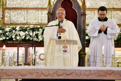 Clérigo de avanzada edad en vestimenta litúrgica blanca habla frente a un micrófono, con joven vestido de túnica blanca a su lado.