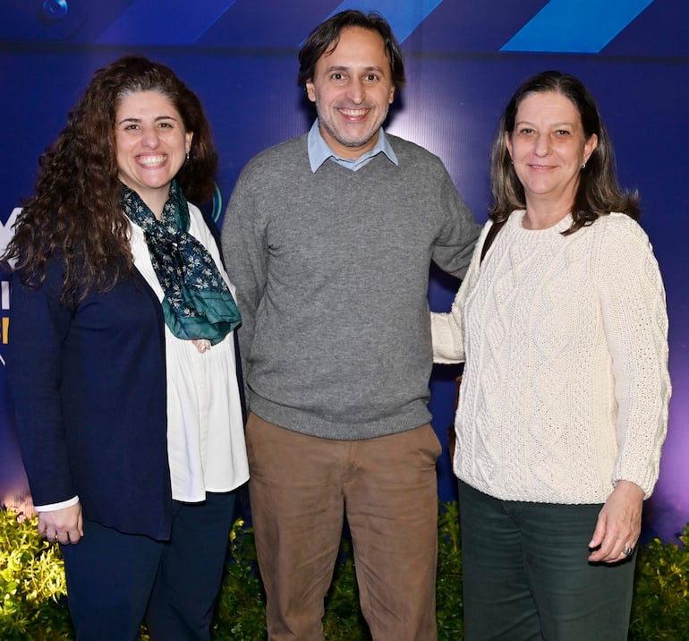 Tres personas sonrientes en evento social: mujer con chal oscuro, hombre con suéter gris, y mujer en suéter blanco, rodeados de decoración azul.