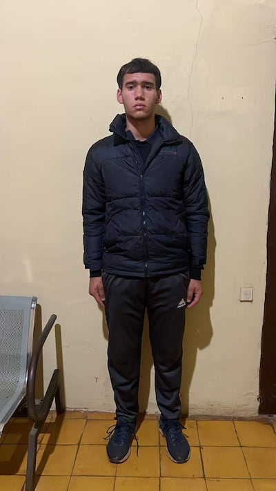Joven de pie en habitación, vestido con abrigo oscuro y pantalones deportivos, mirando a la cámara con atención.