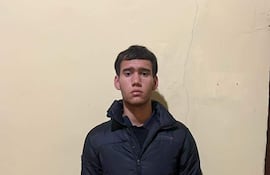 Joven de pie en habitación, vestido con abrigo oscuro y pantalones deportivos, mirando a la cámara con atención.