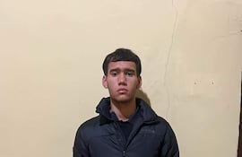 Joven de pie en habitación, vestido con abrigo oscuro y pantalones deportivos, mirando a la cámara con atención.