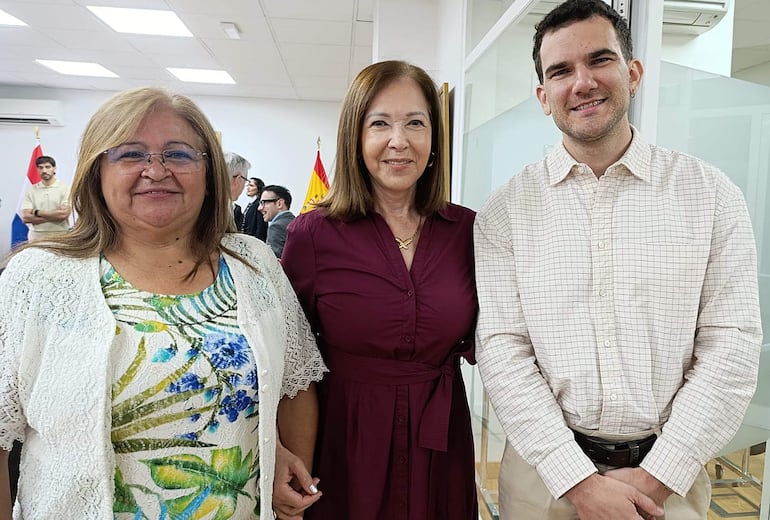 Tres personas: mujer rubia con blusa colorida y abrigo blanco, mujer castaña en blusa burdeos, hombre con camisa a cuadros, todos sonrientes en oficina.