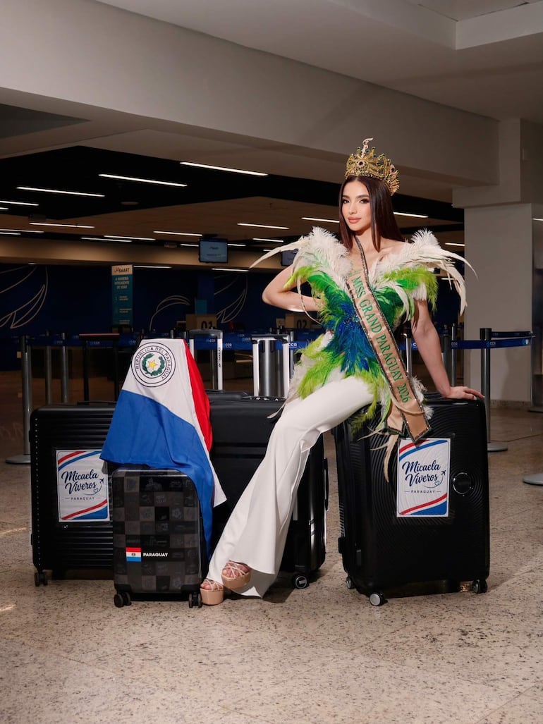 Micaela Viveros con vestido colorido y corona, sentada sobre maletas en el aeropuerto, rodeada de objetos relacionados con Paraguay.