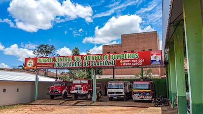 Cartel del Cuartel de Bomberos de Caaguazú con vehículos de emergencias rojos y blancos estacionados bajo un cielo despejado.
