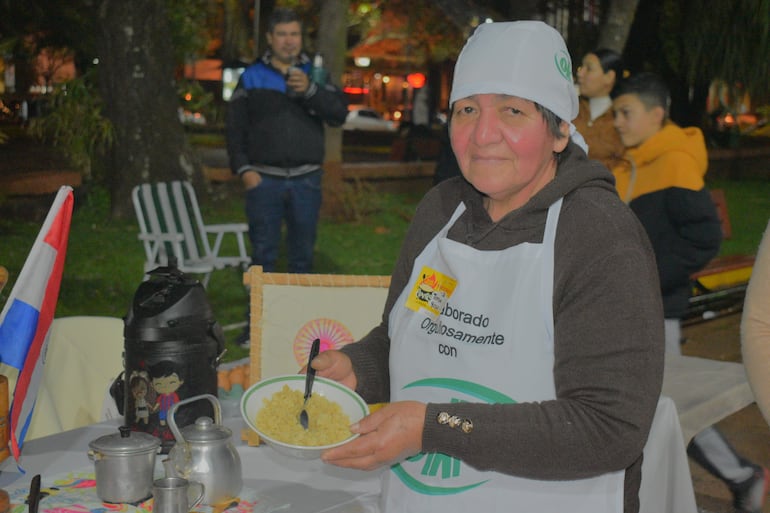 Mujer con gorra blanca y delantal sostiene un plato de comida, rodeada de personas en un ambiente festivo nocturno.