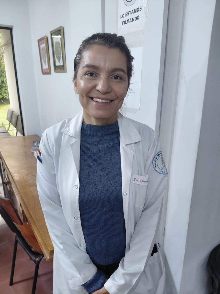 Rossana González sonríe con bata blanca y camiseta azul en un consultorio con sillones de plástico y muebles de madera.