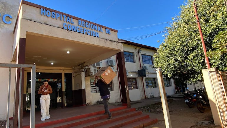 Mujer de pie con teléfono móvil y hombre llevando una caja en la entrada del Hospital Regional de Concepción bajo un cielo despejado.