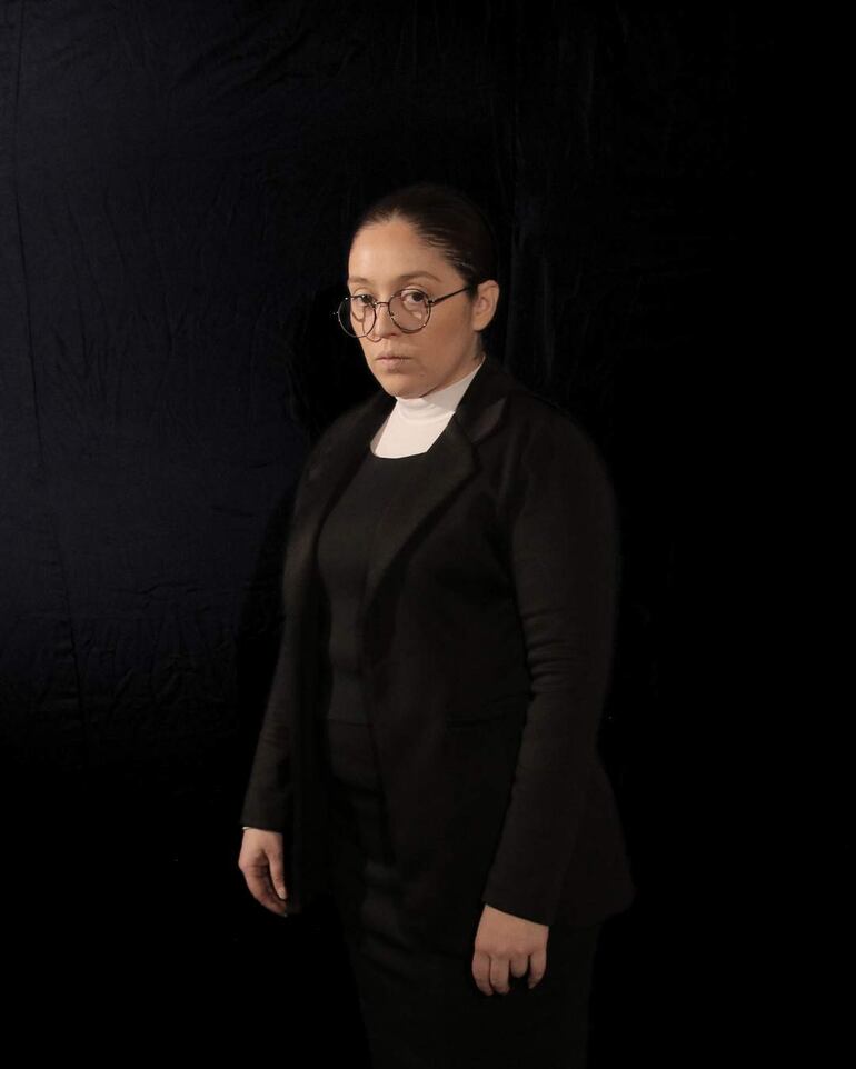 Mujer de pie en un entorno oscuro, con blazer negro y blusa blanca, gafas grandes, expresión seria y postura de tres cuartos.