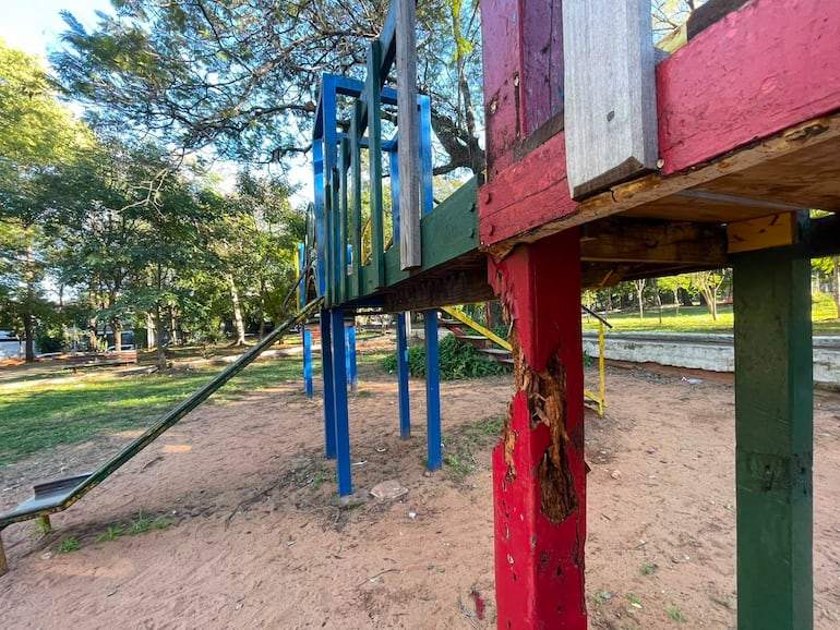 Estructura de juego multicolor deteriorada en un parque vacío, rodeada de césped y árboles.