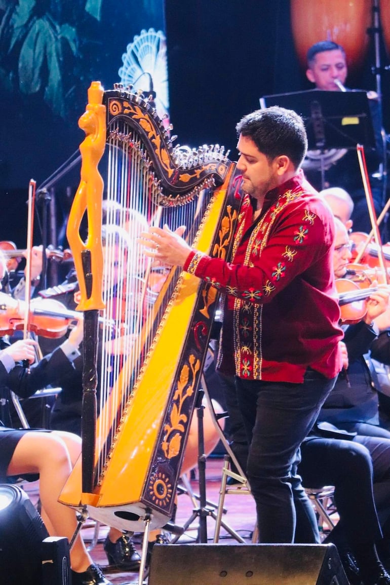 Marcelo Ojeda, con camisa roja y bordados, toca el arpa en el escenario iluminado, rodeado de músicos.