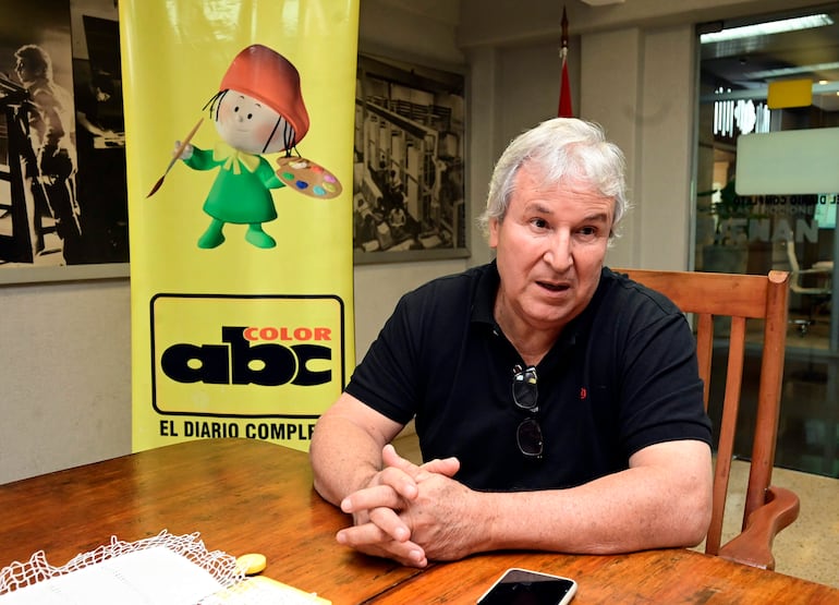 Hombre canoso con camiseta negra y gafas, sentado en mesa de madera, conversando en ambiente de oficina con logo de ABC Color.