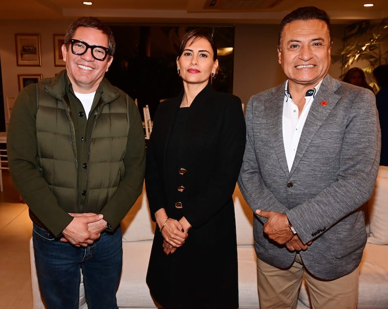 Tres personas sonriendo en evento social: mujer con chaqueta verde y gafas, hombre serio en abrigo negro, y hombre sonriente en blazer gris.