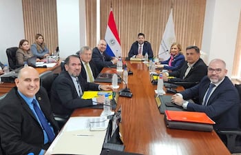 Gerardo Bobadilla sonriente en traje oscuro, rodeado de colegas formales discutiendo en una mesa con documentos y banderas al fondo.
