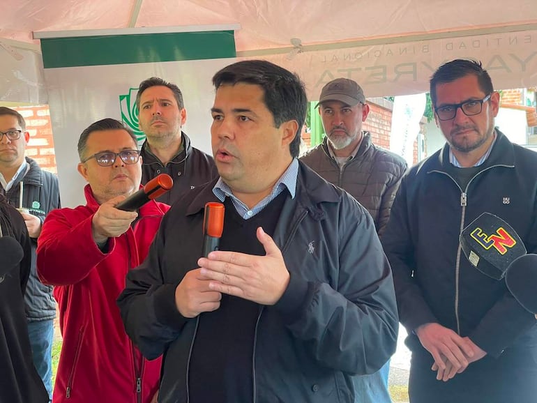 Hombre con micrófono habla en un evento bajo carpa, rodeado de otros cuatro hombres atentos, algunos con micrófonos.