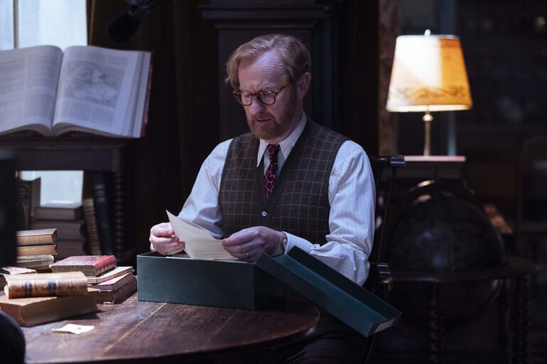 Mark Gatiss sentado en mesa, con chaleco de patrones y corbata burdeos, sostiene un documento rodeado de libros y una lámpara cálida.